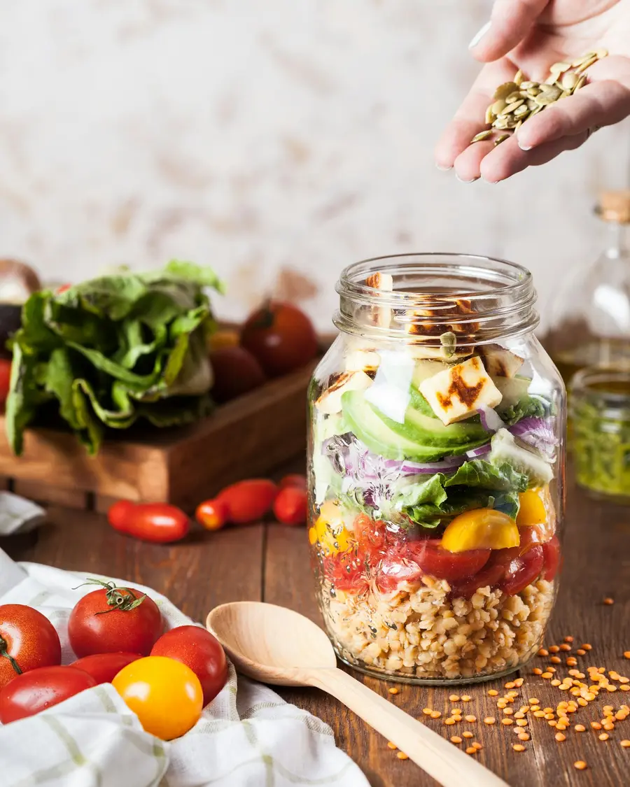 Hand sprinkling seeds over a fresh salad in a glass jar on a sunlit wooden counter
